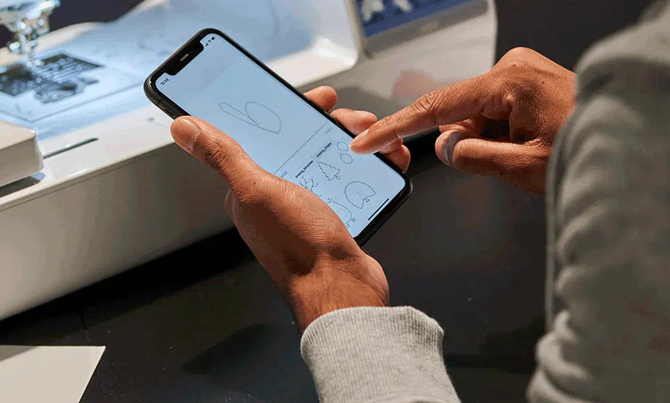 Person using a mobile phone to navigate a sewing and embroidery app, with the Brother Innov-is M380D machine in the background.