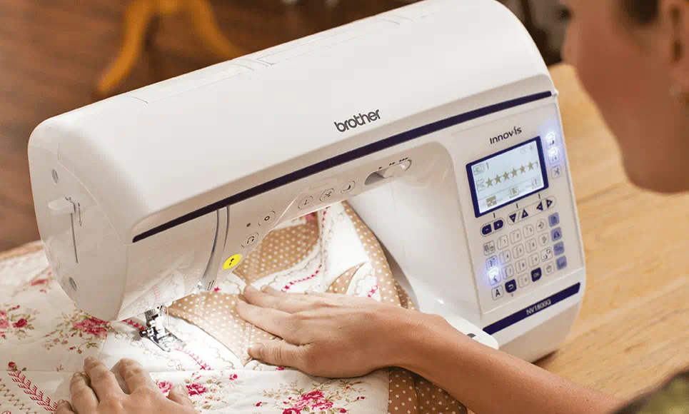 Close-up of a crafter using the Brother Innov-is NV1800Q sewing and quilting machine, highlighting the LCD screen with stitch options and the machine's needle area threading a floral quilt.
